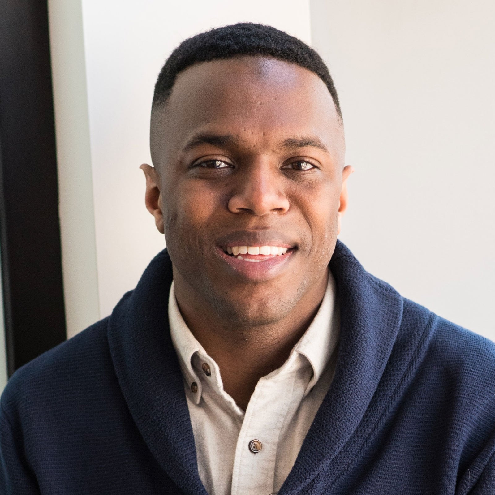 Close-up portrait of a smiling man wearing a beige button-down and navy cardigan indoors.