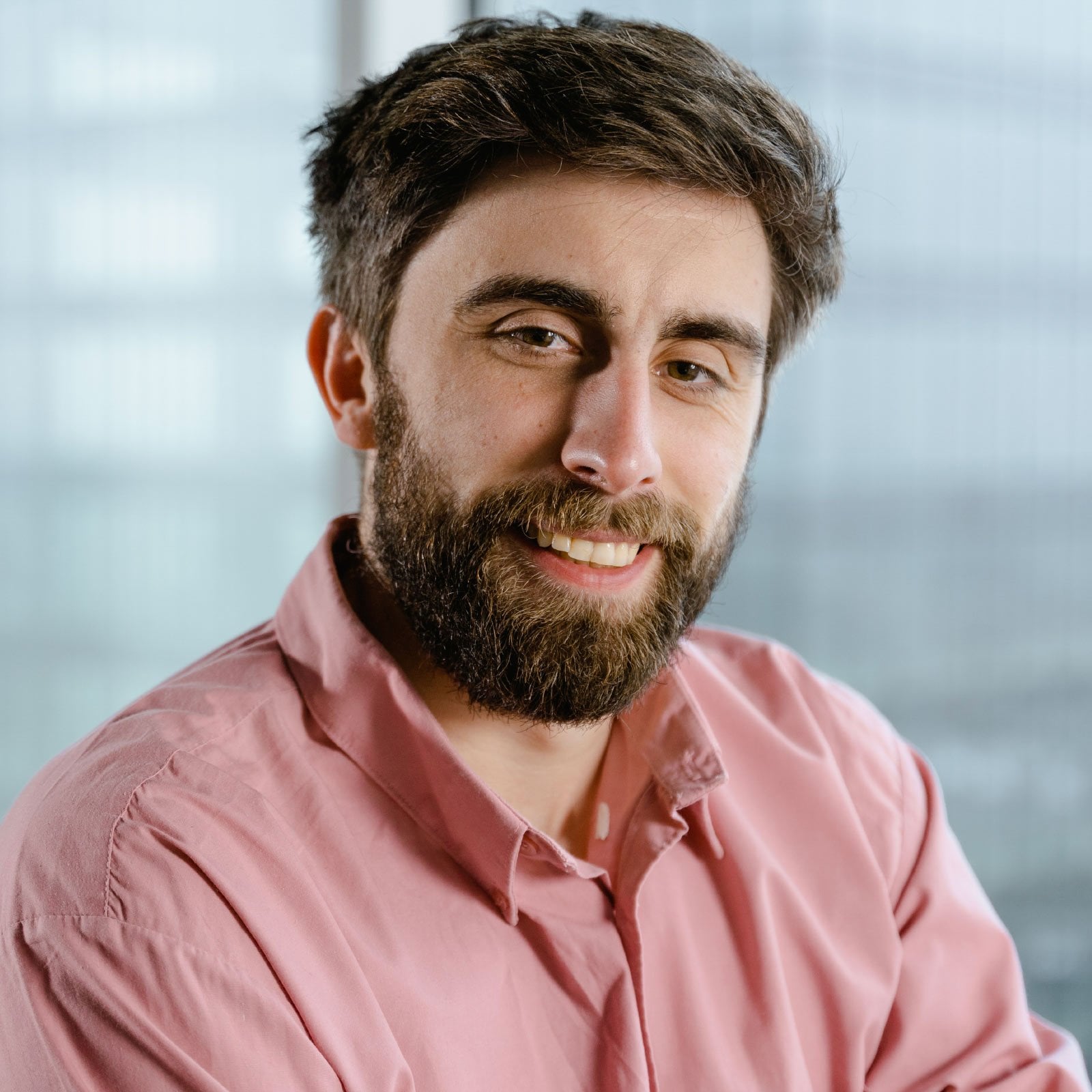 Close-up portrait of a smiling man with a beard, wearing a pink shirt, against a soft office-like background.